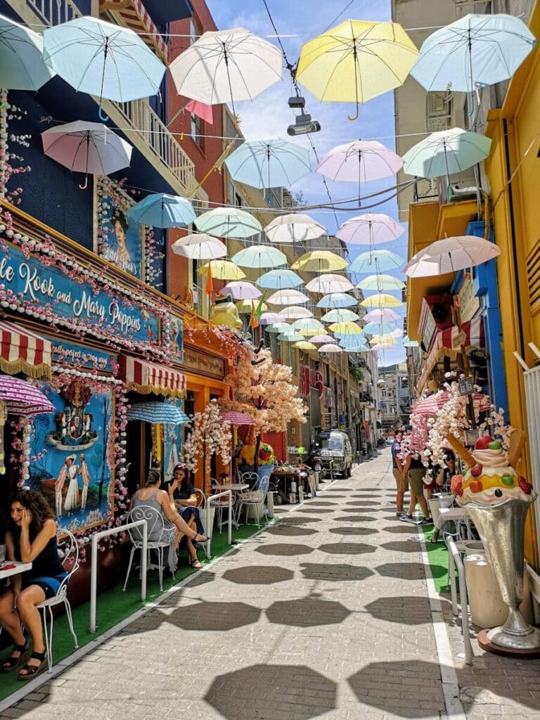 colourful street in Athens with pastel umbrellas overhead and outdoor cafés and shops
