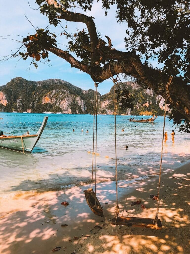 wooden swing hanging from a tree on a Thai beach with longtail boat and limestone cliffs