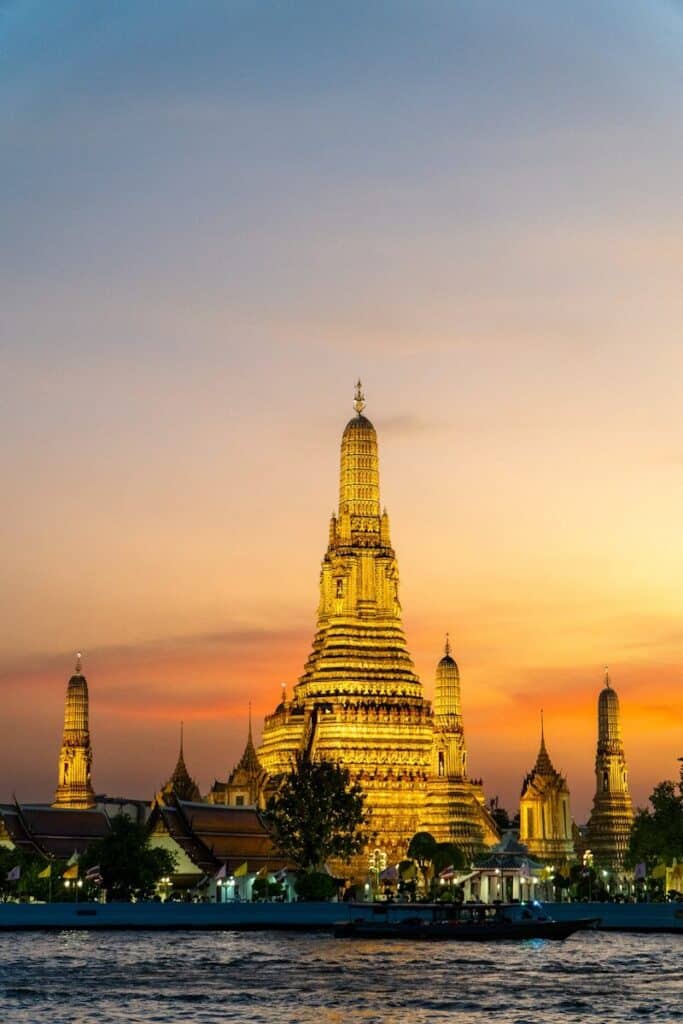 Wat Arun temple in Bangkok illuminated at sunset with orange sky reflecting on the river